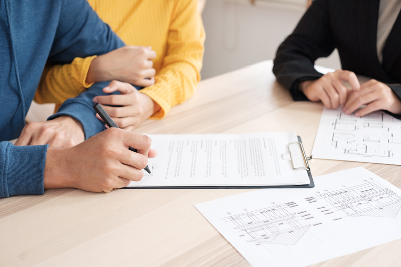 three people are pictured two are signing a contract with while the other is dressed in a suit and appears to be a relator or broker