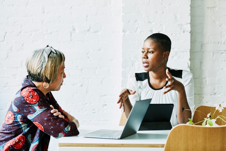 two women seated at a table are speaking to each other with an open laptop in front of one of them