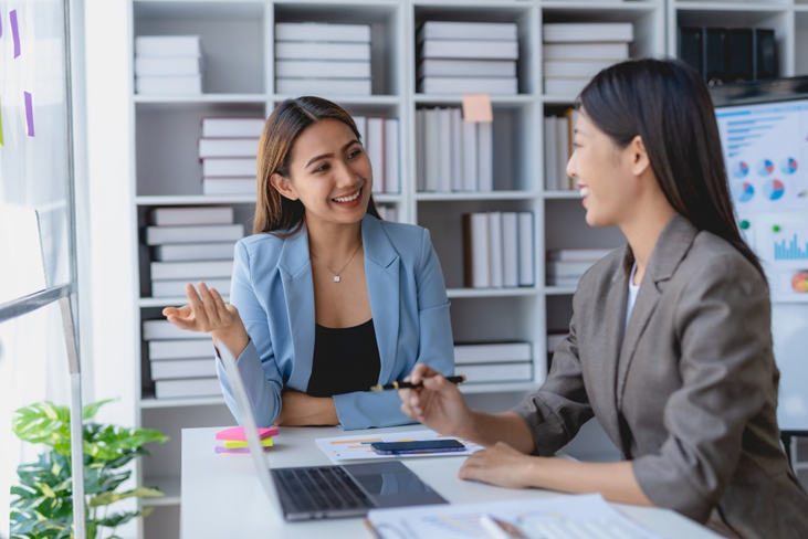 Two businesswomen have a conversation at a conference table in a brightly lit room with white brick walls.
