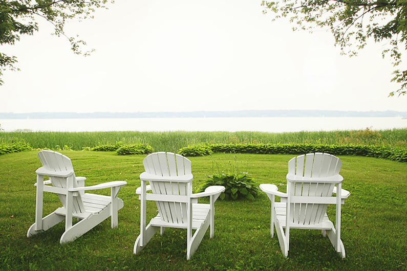 Three white Adirondak chairs are placed in lush green grass overlooking a clear lake.