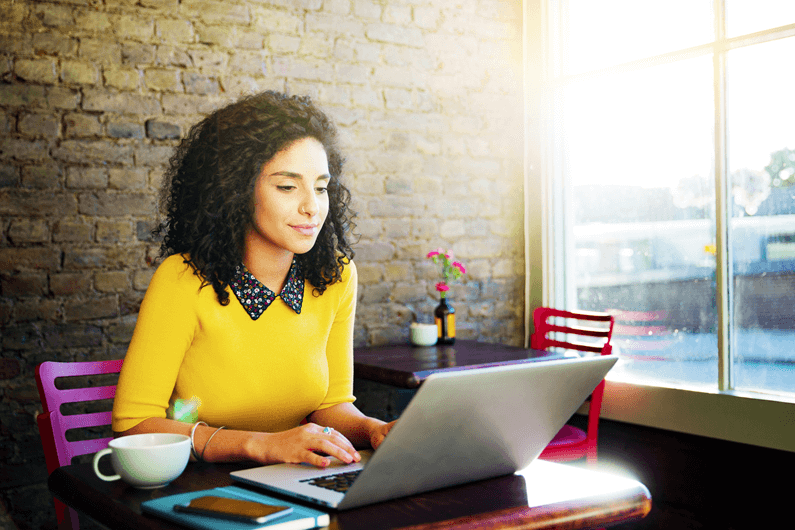 a woman in a yellow sweater smiles at her laptop working in a coffee shop