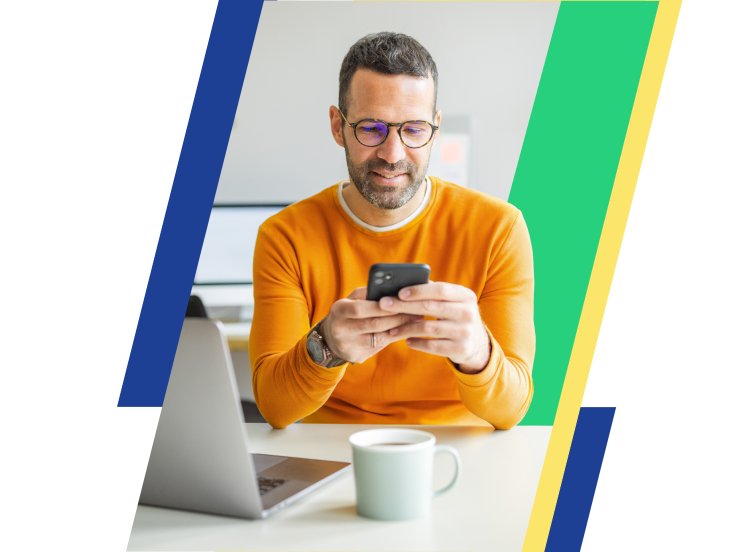 a man in glasses and an orange sweater smiles at a phone in his hands while sitting in front of a laptop and coffee mug