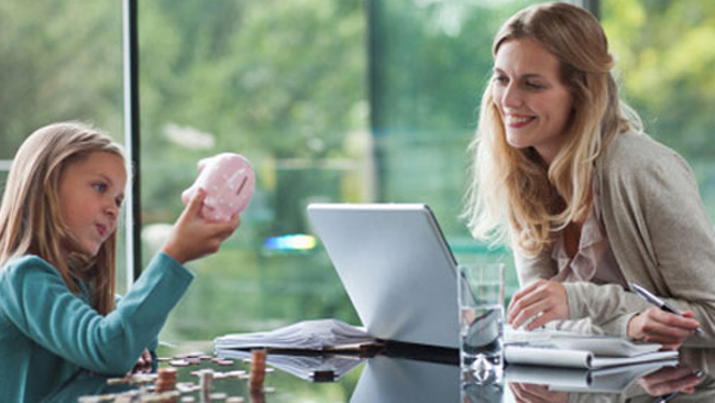 a mom looking at a laptop with a child holding a piggy bank